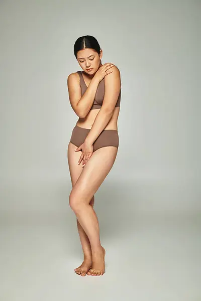 Upset asian woman in underwear covering body while embracing herself on grey backdrop, body shaming — Stock Photo