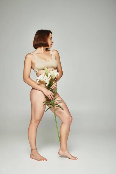Tattooed woman with short hair holding flowers and posing in underwear on grey background, lilies — Stock Photo