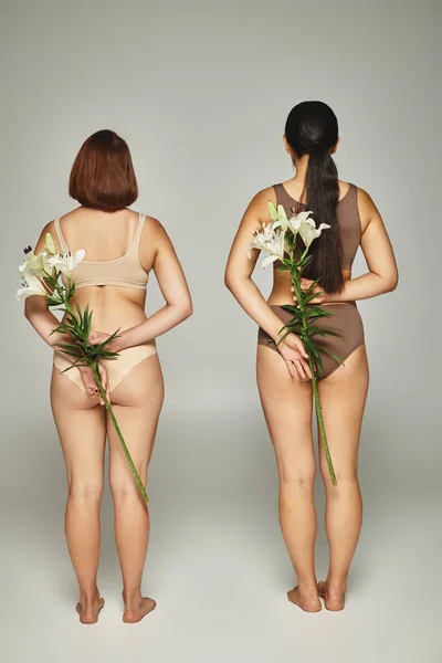 Back view of two women in beige lingerie standing with white lilies in hands on grey backdrop — Stock Photo