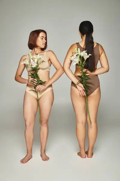 Young woman in beige lingerie standing near female friend and holding flowers in hands on grey — Stock Photo