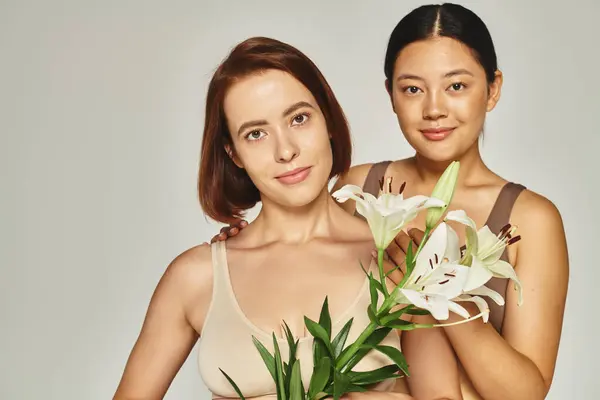 Porträt multikultureller Frauen in Unterwäsche, die zusammen mit Blumen auf grauem Hintergrund stehen — Stockfoto