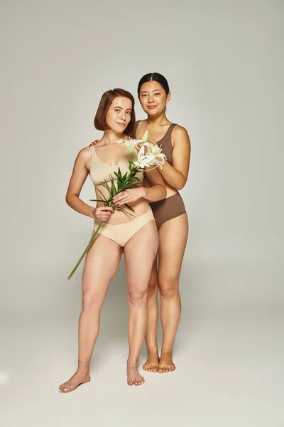 Junge interrassische Frauen in Unterwäsche stehen zusammen mit Blumen auf grauem Hintergrund, in voller Länge — Stockfoto