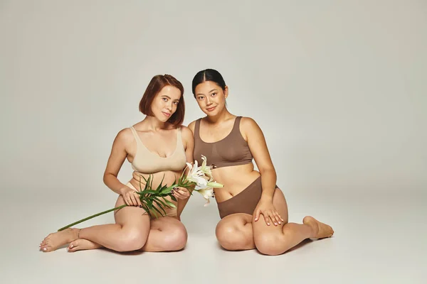 Junge multikulturelle Frauen in Unterwäsche sitzen zusammen mit Blumen auf grauem Hintergrund, Lilien — Stockfoto