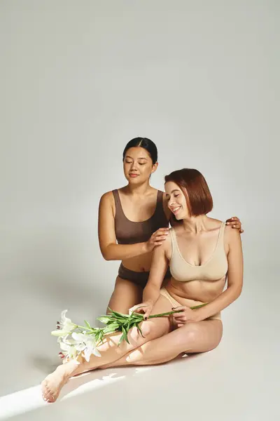 Glückliche multikulturelle Frauen in Unterwäsche sitzen zusammen mit Blumen auf grauem Hintergrund, Lilien — Stockfoto