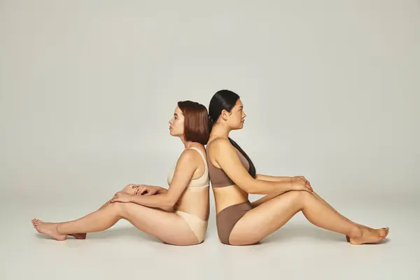 Dos amigas diversas en ropa interior beige y marrón sentadas espalda con espalda sobre fondo gris - foto de stock