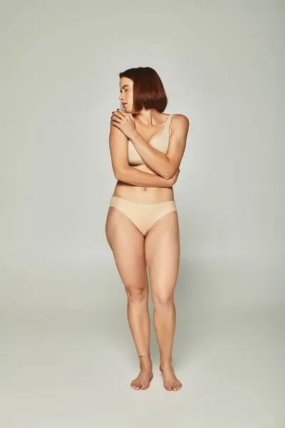 Pleine longueur de femme timide en sous-vêtements beige regardant par-dessus l'épaule sur fond gris, vulnérabilité — Photo de stock