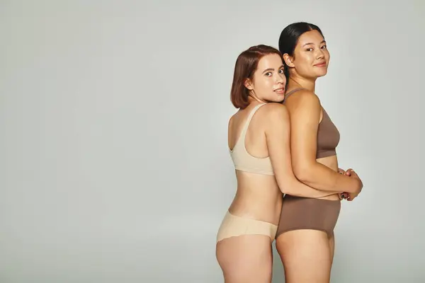 Jóvenes mujeres multiculturales en ropa interior pastel abrazando y mirando a la cámara sobre fondo gris — Stock Photo