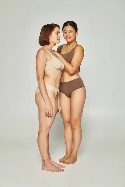 Cheerful asian woman in underwear embracing her friend on grey background, beauty and body positive — Stock Photo