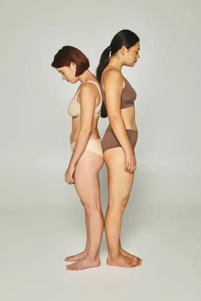 Upset multicultural women in underwear standing back to back on grey backdrop, body shaming — Stock Photo