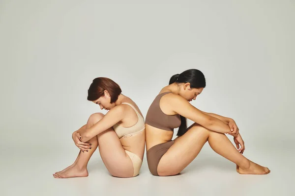Upset multicultural women in underwear sitting back to back on grey backdrop, body shaming — Stock Photo