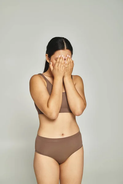 Brunette embarrassed woman in underwear covering face with hands on grey background, body shaming — Stock Photo