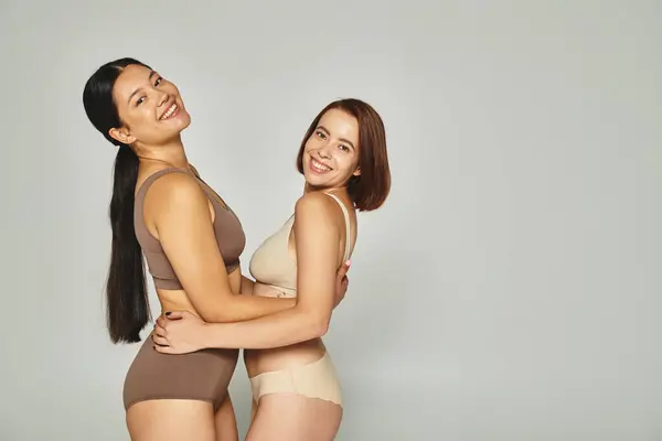 Happy young multicultural women in underwear embracing each other on grey background, body positive — Stock Photo