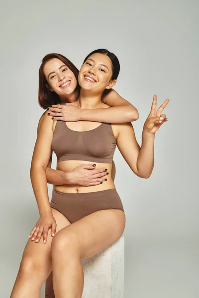 Mulher feliz abraçando seu amigo asiático que mostrando v sinal no fundo cinza, corpo positivo — Fotografia de Stock