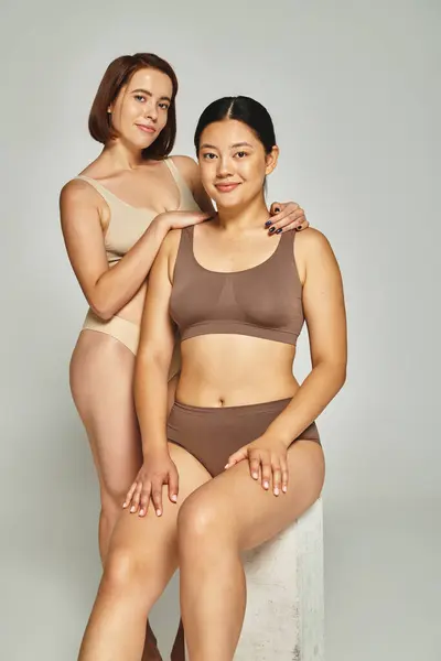 Fröhliche Frau umarmt brünette asiatische Freundin in Unterwäsche auf grauem Hintergrund, Körper positiv — Stockfoto