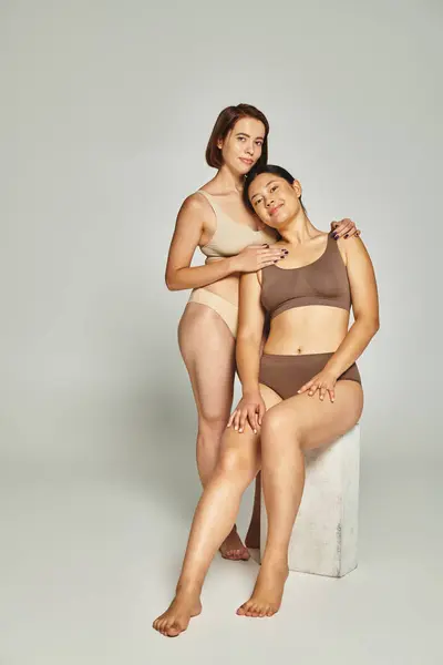 Happy woman embracing brunette asian friend in underwear on grey background, body positive — Stock Photo