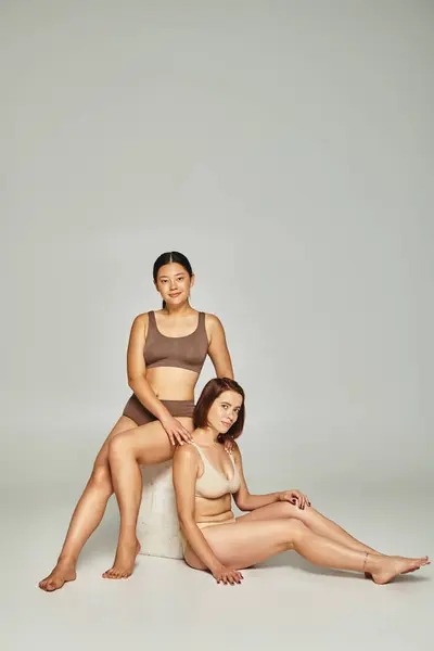 Full length of interracial women in pastel color underwear posing together on grey backdrop — Stock Photo