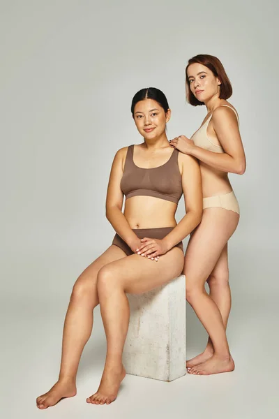 Longitud completa de las mujeres multiculturales en ropa interior de color pastel posando juntos sobre fondo gris - foto de stock