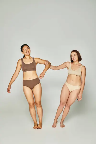 Young woman holding hand on asian female friend in underwear while posing together on grey backdrop — Stock Photo