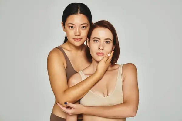 Young asian woman embracing with her female friend in underwear while posing on grey backdrop — Stock Photo