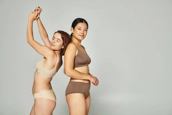 Happy woman in underwear dancing near asian friend in underwear on grey background, body positive — Stock Photo