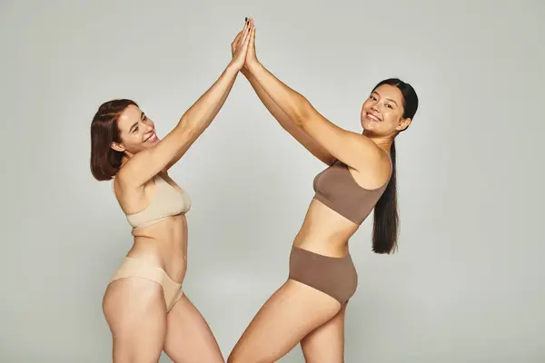 Молодые счастливые межрасовые женщины в нижнем белье дают друг другу пять на сером фоне, тело позитивное — стоковое фото
