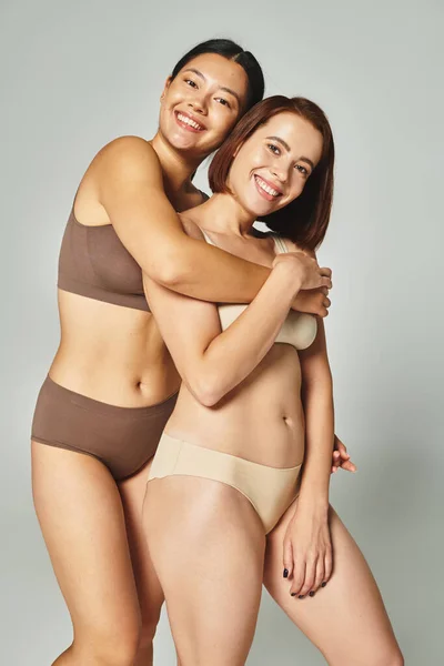 Happy asian woman in underwear embracing her young friend on grey background, companionship — Stock Photo