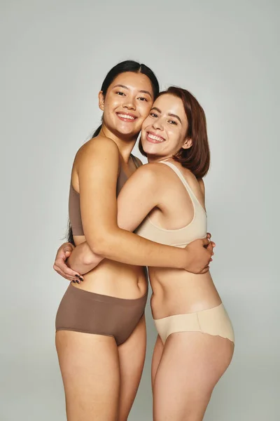 Freudige asiatische Frau in Unterwäsche umarmt ihre Freundin auf grauem Hintergrund, Freundschaft — Stockfoto