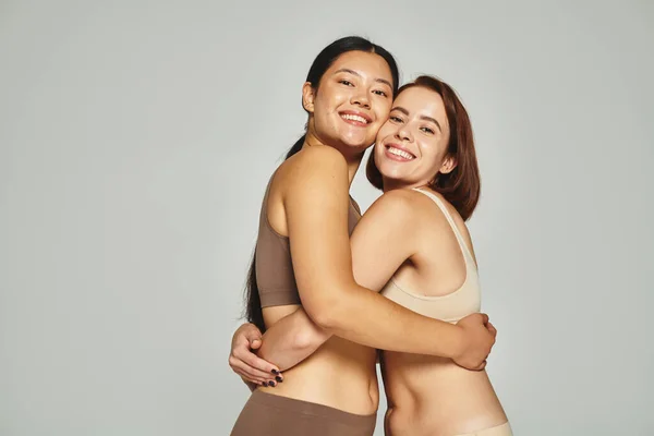 Mujeres alegres y jóvenes multiétnicas en ropa interior de color pastel abrazando sobre fondo gris - foto de stock