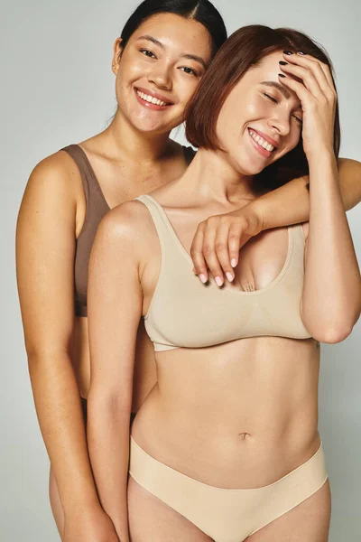Femmes gaies et multiethniques en sous-vêtements de couleur pastel embrassant et souriant sur fond gris — Photo de stock
