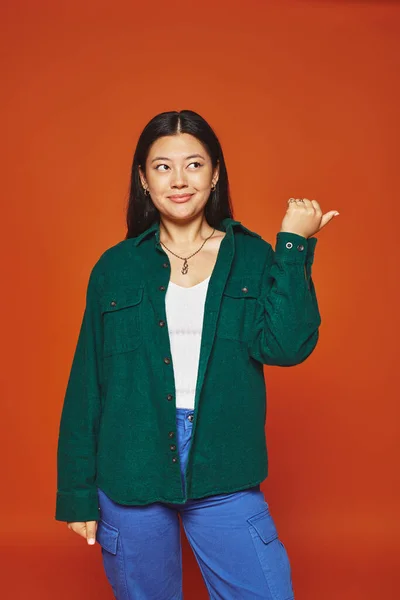 Asiatin posiert in lebendigem Outfit mit grüner Jacke, die mit Daumen auf orangefarbenem Hintergrund zeigt — Stockfoto