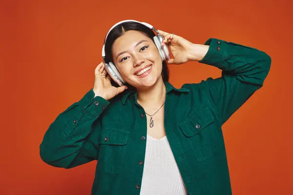 Young and brunette asian woman enjoying music with wireless headphones on orange background — Stock Photo