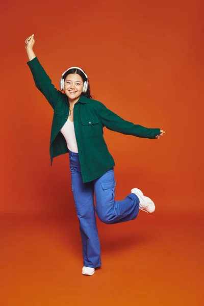 Full length of brunette asian woman enjoying music with wireless headphones on orange background — Stock Photo
