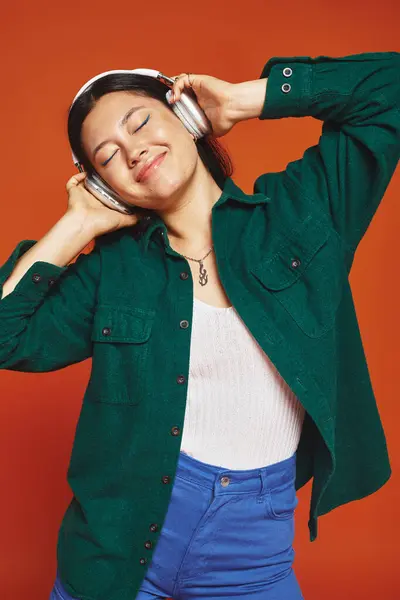 Pleased brunette asian woman enjoying music with wireless headphones on orange background — Stock Photo