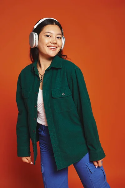 Sorridente bruna donna asiatica godendo della musica con cuffie wireless su sfondo arancione — Foto stock
