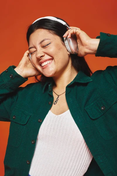 Positiva e morena mulher asiática desfrutando de música com fones de ouvido sem fio no fundo laranja — Fotografia de Stock