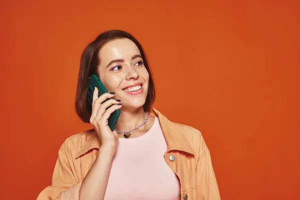 Happy young woman in vibrant attire talking on smartphone on orange background, phone call — Stock Photo