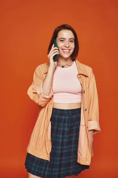 Cheerful young woman in vibrant attire talking on smartphone on orange background, phone call — Stock Photo