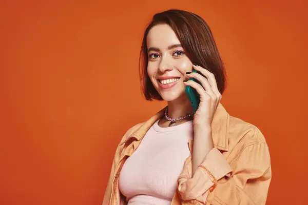 Jovem mulher positiva em traje elegante falando no smartphone no fundo laranja, chamada telefônica — Fotografia de Stock