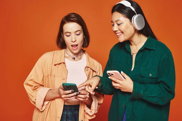 Dos alegres amigos multiculturales usando smartphones y escuchando música sobre fondo naranja - foto de stock
