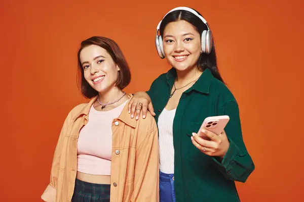 Glückliche Asiatin mit drahtlosen Kopfhörern, die ihr Smartphone in der Nähe ihrer Freundin in Orange hält — Stockfoto