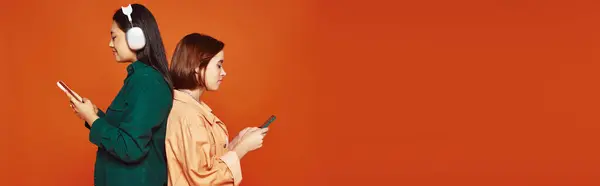 Glückliches asiatisches Mädchen in drahtlosen Kopfhörern mit Smartphone in der Nähe ihrer Freundin auf orangefarbenem Banner — Stockfoto