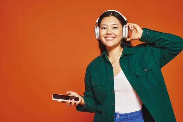 Happy asian woman listening music in wireless headphones holding smartphone on orange background — Stock Photo
