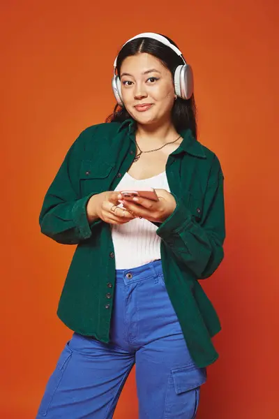 Cheerful asian woman listening music in wireless headphones holding smartphone on orange background — Stock Photo
