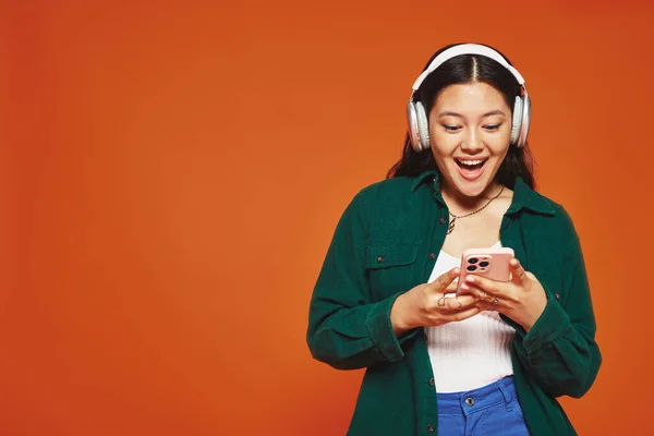 Excited asian woman listening music in wireless headphones using smartphone on orange background — Stock Photo