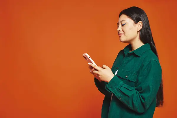 Joyful and young asian woman with brunette hair using smartphone and smiling on orange background — Stock Photo