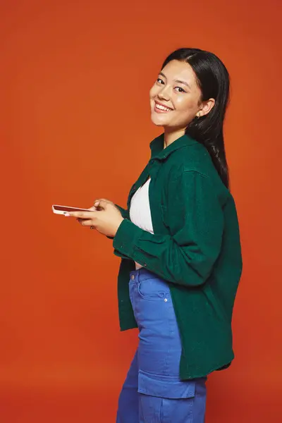 Fröhliche und junge asiatische Frau mit brünetten Haaren mit Smartphone und Lächeln auf orangefarbenem Hintergrund — Stockfoto