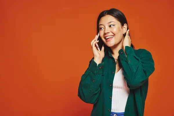 Cheerful young asian woman with brunette hair talking on smartphone and smiling on orange background — Stock Photo