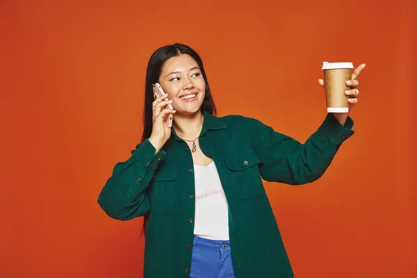 Happy asian woman talking on smartphone and holding paper cup with coffee on orange background — Stock Photo