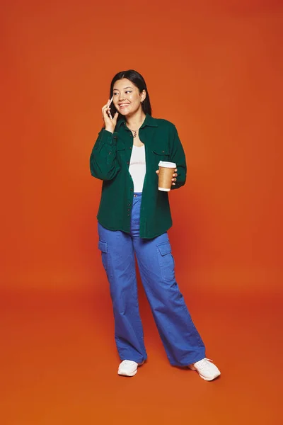 Alegre mulher asiática falando no smartphone e segurando copo de papel com café no fundo laranja — Fotografia de Stock