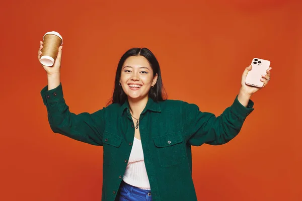 Excité asiatique femme parler sur smartphone et tenant tasse en papier avec café sur fond orange — Photo de stock